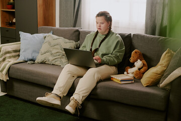 Young female student with Down syndrome wearing casual clothes sitting on couch in living room,...