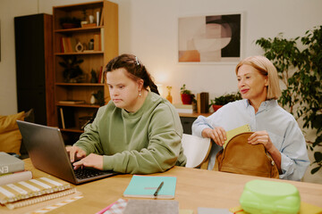 Mature woman taking out notebook from backpack, while her daughter with Down syndrome typing on...