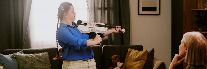 Modern girl with Down syndrome standing in living room, playing violin in front of her proud...