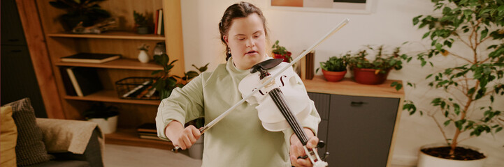 High angle view of young woman with disability standing in cozy living room, practicing violin with focused expression, header for website
