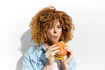 Young woman with curly hair enjoying a delicious burger against a minimalist background, showcasing joy and indulgence in fast food culture