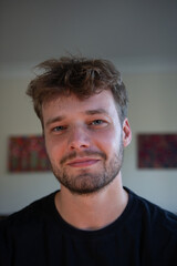 A close-up indoor portrait of a man with a calm and relaxed expression, soft lighting accentuating his features. The blurred background with colorful artwork adds warmth and depth to the image