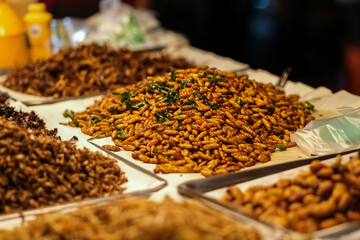 Close-up view of a traditional Thai street market stand showcasing trays of fried insects, including crickets and beetles.