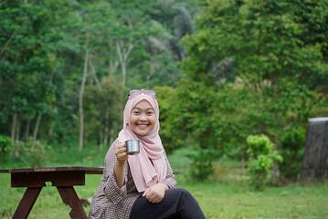 Young Indonesian woman wearing hijab enjoying the calming natural atmosphere while enjoying a cup of black coffee, away from the hustle and bustle of the city, travel concept.