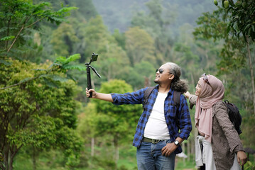 Fototapeta premium Indonesian man with curly hair and young Indonesian woman wearing hijab taking vlog video while walking in the forest while enjoying the beauty of natural scenery, travel concept.