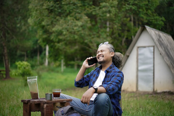 Fototapeta premium Indonesian man with curly hair is on a voice call while enjoying a cup of kopi tubruk while camping in a forest cabin, travel concept.