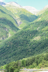 Fototapeta premium Norwegian mountain landscape with green trees casting shadows. White snow on the top of the fjords under the blue sky of a sunny summer day. A road at the foot of the mountain.