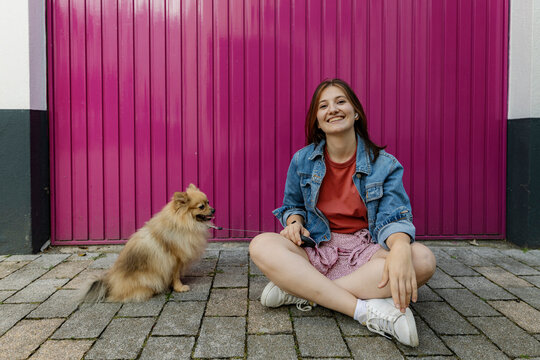 Happy woman sitting with Pomeranian dog in front of magenta wall
