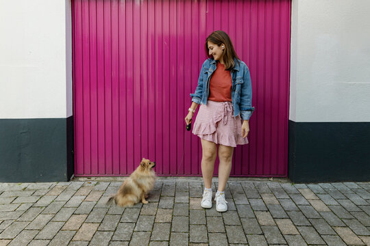Happy woman standing near Pomeranian dog in front of magenta wall