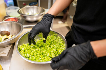 A chef wearing black gloves prepares diced green vegetables in a metal bowl, surrounded by other ingredients in a bustling kitchen setting. Freshness and culinary skills are highlighted.