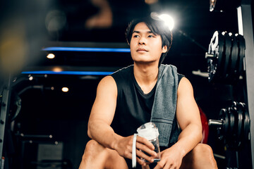 Portrait of Asian young man trained body in black sportswear sitting for rest holding water or protine in fitness gym, Weight training, healthy lifestyle concept