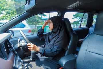 A woman wearing a black hijab is driving a car. She is looking at the steering wheel. The car is...