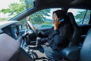 A woman is talking on her cell phone while driving a car. She is wearing a black hijab and is driving a white car