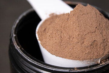 Close up of a scoop full with chocolate protein powder on a black can, grey table background