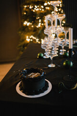 Festive black cake with Happy Birthday inscription. Blowing out candle and making a wish. Glasses at a champagne party on Christmas night against a background of a Christmas tree with garlands