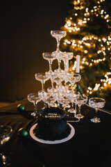 Festive black cake with Happy Birthday inscription. Blowing out candle and making a wish. Glasses at a champagne party on Christmas night against a background of a Christmas tree with garlands