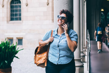 Fototapeta premium Cheerful woman in casual clothing and trendy sunglasses laughing during friendly cellphone conversation, happy female student with backpack calling via mobile application during break at university