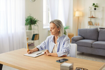 Senior woman working from home, writing a business and finance plan in notepad. Focused and positive approach demonstrates dedication to planning and productivity in a personal workspace.