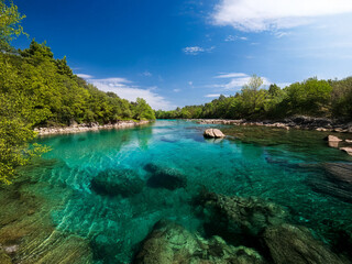 Fototapeta premium Serene river scene with crystal-clear turquoise water, lush green banks, and a bright blue sky. Rocks are visible beneath the surface. The tranquil atmosphere invites relaxation and exploration.