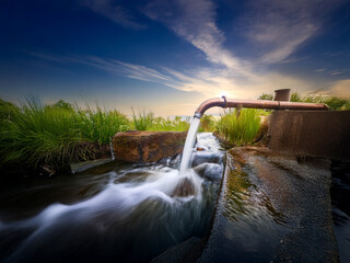 Serene sunset scene featuring a copper pipe dispensing fresh water into a rocky stream.  The water flows smoothly over the stones, creating a tranquil atmosphere.