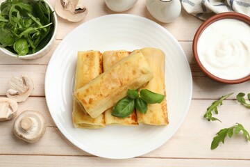 Delicious rolled crepes with basil, arugula, mushrooms and sour cream on wooden table, flat lay