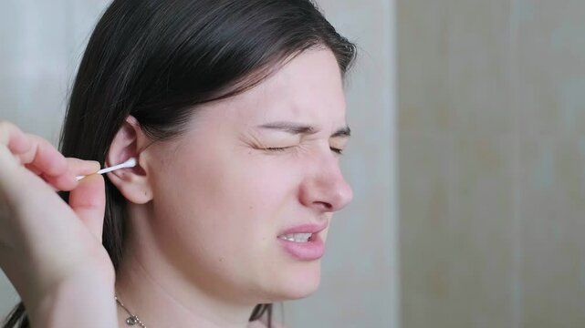 Millennial woman cleaning her ears in the bathroom, feeling pain during ear cleaning and making a grimacing facial expression.