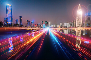 Fototapeta premium Night scene of vibrant Shenzhen, China. A long exposure captures streaks of light from speeding vehicles on a highway, reflecting on calm waters, with a modern cityscape backdrop.