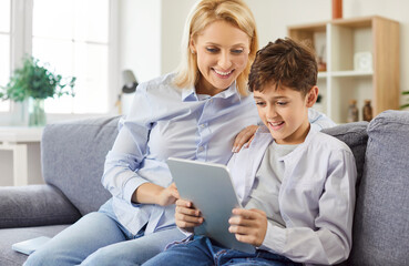 Portrait of a happy young cheerful mother using tablet with her little child son sitting on sofa in living room at home watching funny cartoon enjoying weekend. Family leisure and technology concept.