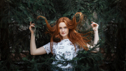 girl with long red hair. She stands against a background of green spruce branches. The girl is wearing a light T-shirt with a floral pattern.