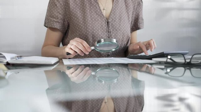 Businesswoman accountant in brawn dotted blouse reviewing financial reports with magnifying glass and calculator, conducting audit and analyzing data in office. Taxes, audit in business