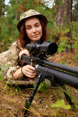 Fototapeta premium A woman with brown hair and a camouflage outfit lies on the forest floor, aiming a rifle at a target. The greenery surrounds her as she concentrates on her training