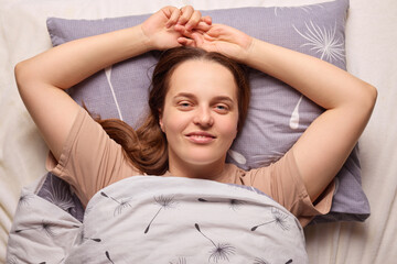 Fototapeta premium A woman lies in bed, her arms raised behind her head, smiling contentedly. The soft bedding and pillow create a cozy atmosphere in the room, capturing a moment of relaxation