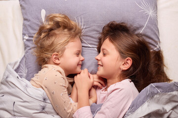 Two young girls lie close together in bed, smiling and looking into each other\'s eyes. They are enjoying a playful moment under soft blankets in a cozy morning setting