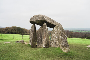 A weathered dolmen with a flat stone atop three upright stones sits in a lush green landscape,...