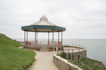 Fototapeta premium The image features a charming seaside bandstand made of metal and wood, situated on a grassy coastal cliff overlooking the ocean. The structure is octagonal with a peaked roof.
