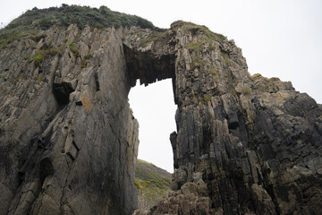 Towering cliffs with a natural arch silhouette against an overcast sky. Rugged gray rocks and sprouting greenery create a dramatic, moody scene, perfect for exploration and inspiration.