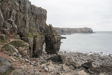 Rugged coastal landscape with towering cliffs, a natural rock arch, and a pebbly shoreline. The overcast sky and gentle ocean waves create a tranquil atmosphere, near St Govan's Chapel. 