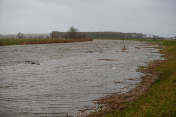 Floodwaters cover a canal,  surrounding  fields and trees under a cloudy sky.  Windmills are visible in the background.