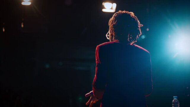 Popular comedian or life coach performing on stage, rearview in soffit lights. Back view of mixed race male person speaking for audience, emotional charismatic guy with dreadlocks, entertainment