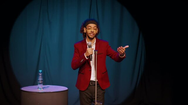 Popular comedian performing on stage, speaking humors and jokes, portrait shot. Handsome young adult African man with microphone standing alone in light of soffit, stand-up comedy concert, medium shot