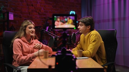 Man host and woman guest recording interview in loft podcast studio using microphones and camera, creating digital content, presenter and woman start conversation.