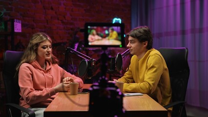 Man host and woman guest recording interview in loft podcast studio using microphones and smartphone on tripod, creating digital content, camera screen pov.