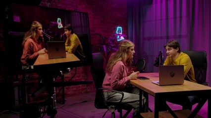 Backstage shot of host and guest recording interview in loft podcast studio, creating digital content, presenter and woman talk with guest on laptop, monitor at side with video stream.