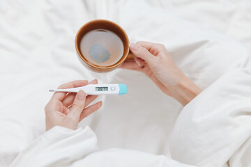 Sick person in bed holding a thermometer and cup of coffee, with another thermometer on the bedside...