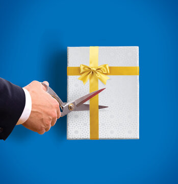 A hand of an executive, poised with scissors, prepares to unveil a beautifully wrapped gift. The scissors are ready to cut the golden ribbon of the gift box. Conceptual photo to depict reward or perk.