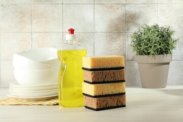 Stack of sponges, detergent and dishware on light wooden table, closeup