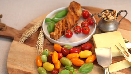 Wooden board with eco-friendly fresh ingredients: multicolored cherry tomatoes, leafy basil, golden cheese slice, walnut bowl, flaky twist pastry. Emphasis on clean, healthy, sustainable food concept.
