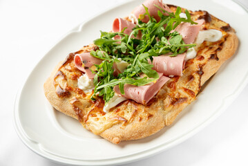 A delicious flatbread pizza topped with slices of ham, fresh arugula, and a creamy sauce. Perfect for food photography and culinary presentations. Ideal for menus and promotional materials.