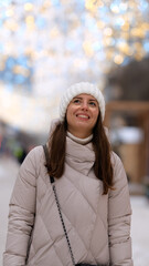 Portrait of a beautiful young woman in winter clothes. The woman smiles happily against the background of a snowy street decorated with Christmas garlands.