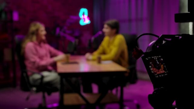 Backstage blurred shot of host and guest recording interview in loft podcast studio, creating digital content, camera on tripod filming at side, display pov. Slow motion.